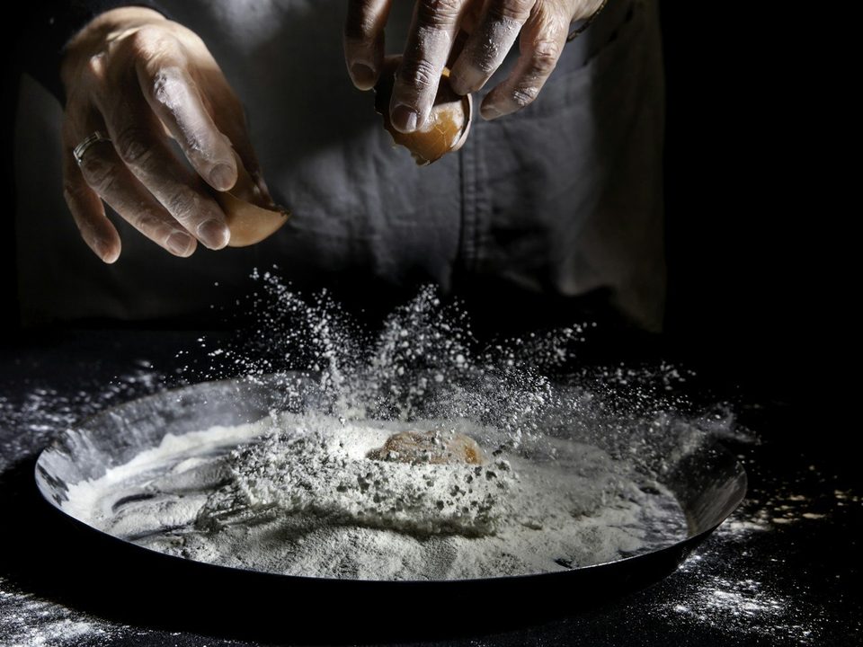 Picture 1: Hands cracking an egg into a metal bowl filled with white flour